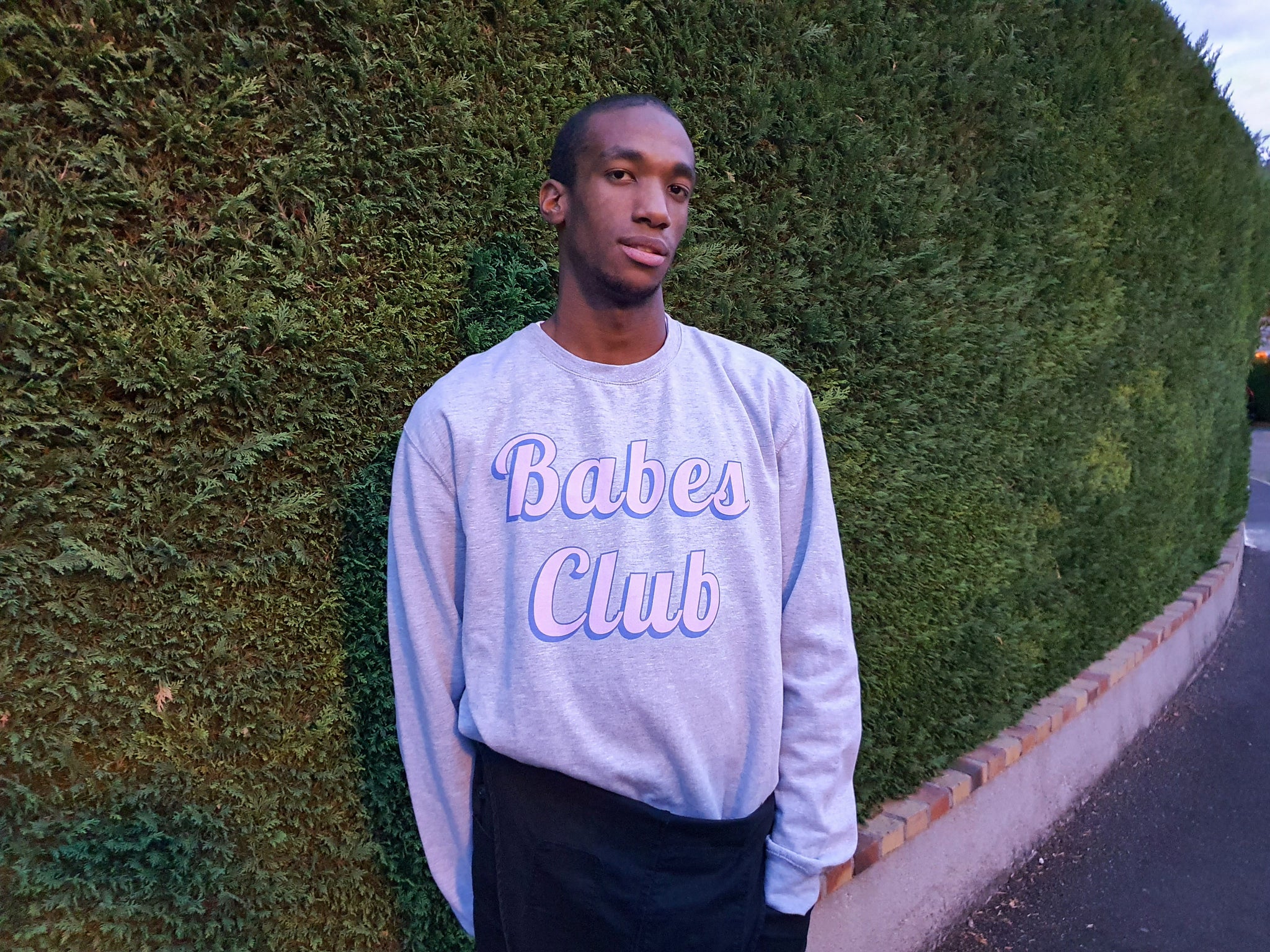 Antoine porte le sweat-shirt baves club devant une belle haie de jardin verte.