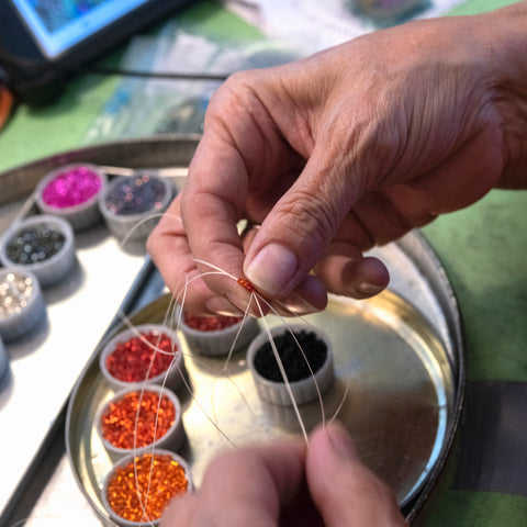 A photograph of two hands stringing an array of tiny, colorful beads onto fishing line.