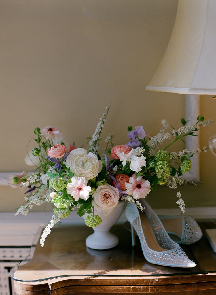 A light and airy spring floral arrangement in blush pink, peaches and lilac is on a table next to a pair of white pointy wedding shoes.