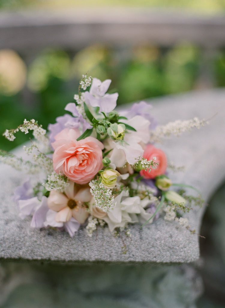 An airy bouquet of spring flowers in peach, lilac and blush pink lies flat on a cement bench