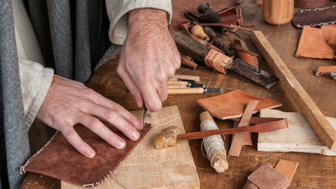 male hands sewing scraps of leather on a workshop bench surrounded by rustic materials