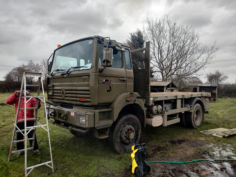 Army green flat bed truck in field with overcast skies, next to a man in a red coat standing on a pair of ladders
