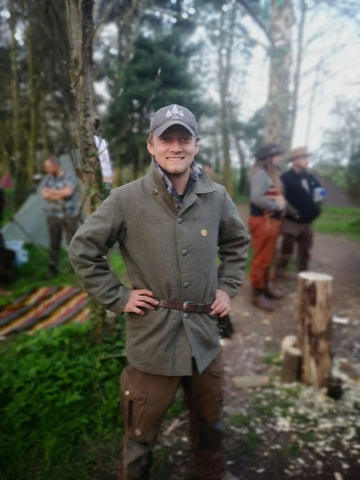 Man stands smiling in forest wearing grey military jacket and surrounded by other outdoor enthusiasts and tents