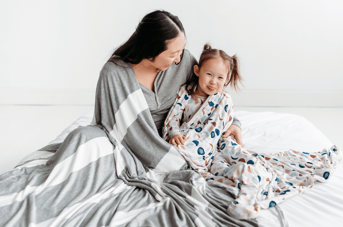 Mother cuddling her daughter with Heather Gray Striped Oversized Cloud Blanket draped around her and her daughter is cuddling with the Luna Neutral Cloud Blanket