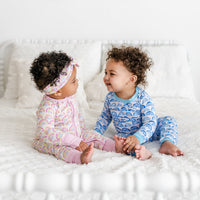 image of baby girl and baby boy sitting on a bed with pink and blue rainbow printed pajamas. Baby boy is pictured wearing blue rainbow printed pajama set, which features a white background and shades of blue rainbows with sky blue trim accents. Baby girl is wearing pastel pink rainbow printed zip up romper with matching rainbow printed bow headband. 