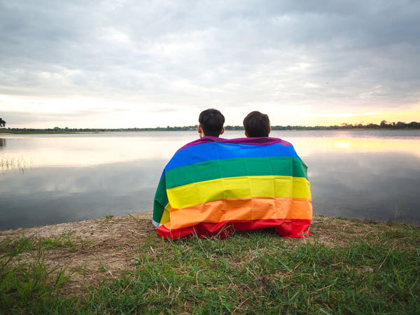 Two men sitting on the edge of a lake with a pride flag draped over their shoulders