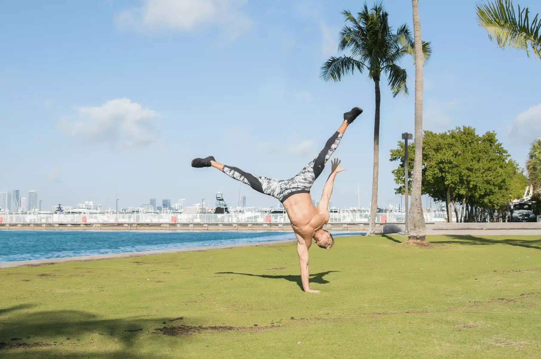 Men's sports leggings in a gray and black pattern, worn during a one-handed handstand outdoors near palm trees and waterfront.