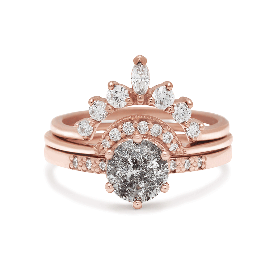 A solitaire ring with a round galaxy grey diamond with white diamond pave on either side set in rose gold set beneath two white diamond bands one with marquise diamonds and a crescent shaped band.