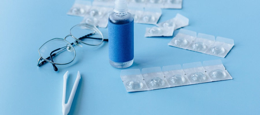 closeup of unused contact lenses in original packaging with lens solution and pair of eyeglasses against light blue background