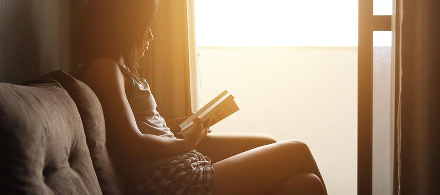 Mujer leyendo un libro en el sofá temprano en la luz del sol visto desde un lado a la luz de la puerta abierta del balcón