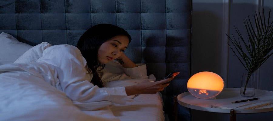 Una mujer abotonando su teléfono en la cama por la noche incapaz de conciliar el sueño con una mesita de noche con una luz naranja junto a ella