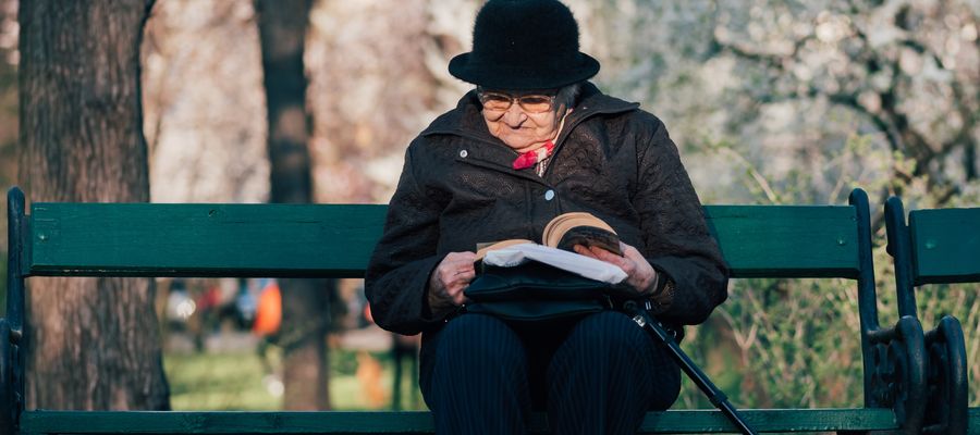 old woman with a hat and eye glasses sitting on a park bench reading with walking cane leaning on her leg