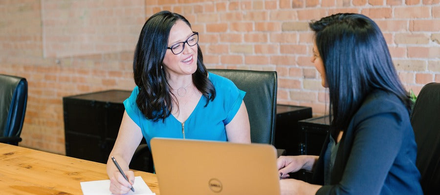 two women with dark hair talking at office desk before computer while the one with glasses smiles and writes something down
