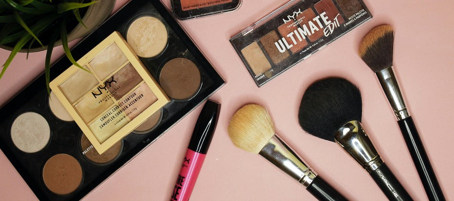 mascara, eye shadow, and makeup brushes scattered on a washed out pink surface seen from above