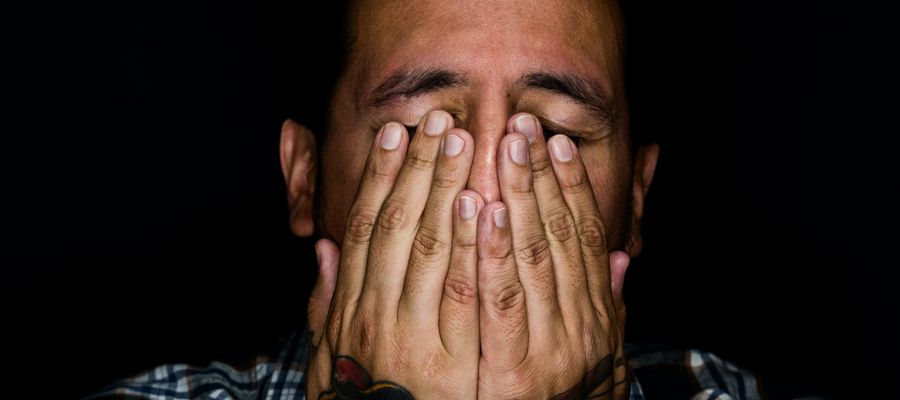 man covering eyes with hands against black background