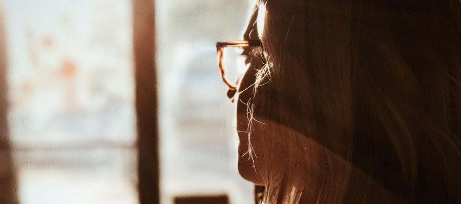Mujer con gafas cubierta de sombra vista de perfil mirando la ventana iluminada por el sol