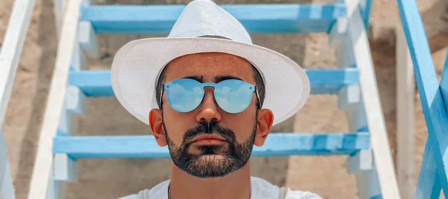 man with short black beard wearing sunglasses and white hat against a background of blue steps