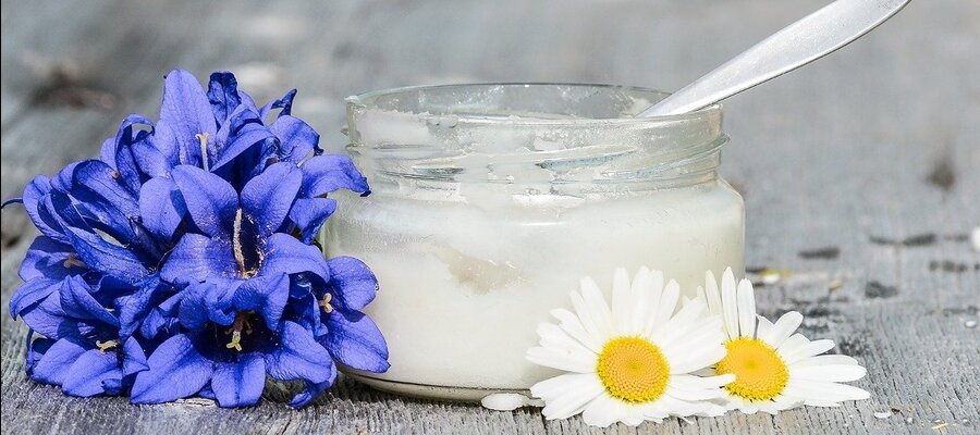 glass jar with virgin coconut oil and silver spoon in it with violet blue flowers to the left and daisies on the right