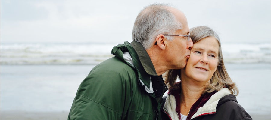 anciano con diabetes y ojos secos besando a su esposa en la mejilla en la playa con olas en el fondo