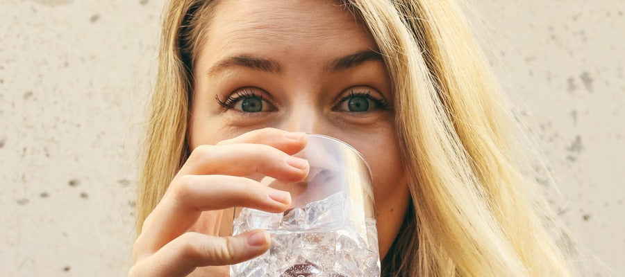 Mujer con cabello rubio bebiendo agua de un vaso transparente con cubitos de hielo y levantando las cejas.