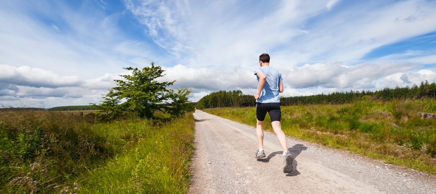 Hombre corriendo por una carretera rural visto desde atrás con un cielo azul lechoso y campos verdes a ambos lados