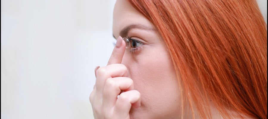 Mujer pelirroja vista de perfil poniéndose lentes de contacto en el ojo izquierdo.