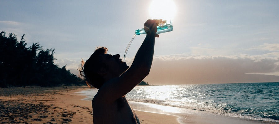 silueta negra de un hombre bebiendo agua en la playa de una botella de plástico que sostiene sobre su cabeza bajo el sol