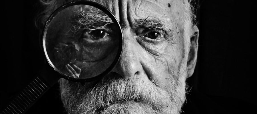 lente grande sostenida sobre el ojo de un anciano con barba y bigote que tiene enfermedad del ojo seco en un retrato en blanco y negro