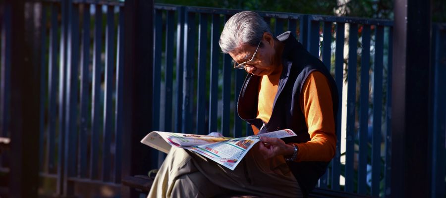 old man wearing glasses sitting on a chair reading a newspaper