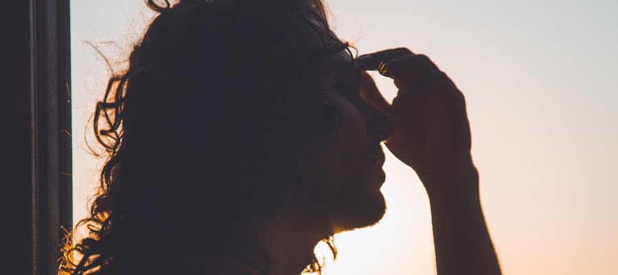 man standing by a window taking hand to forehead because she has a headache