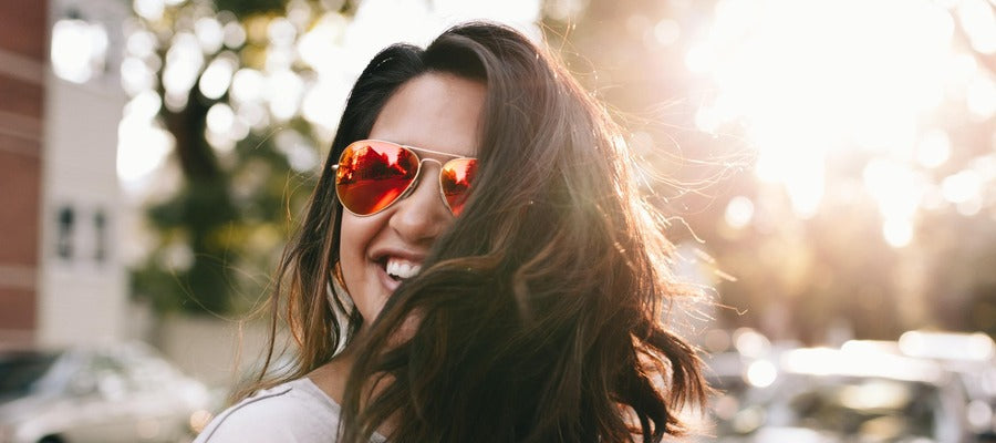 woman with sunglasses and long hair smiling outside against blurry background with her head turned away from the sun