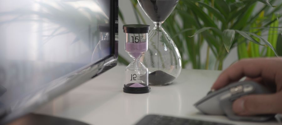 reloj de arena en el escritorio frente a la computadora con una mano sobre un mouse en el borde de la pantalla