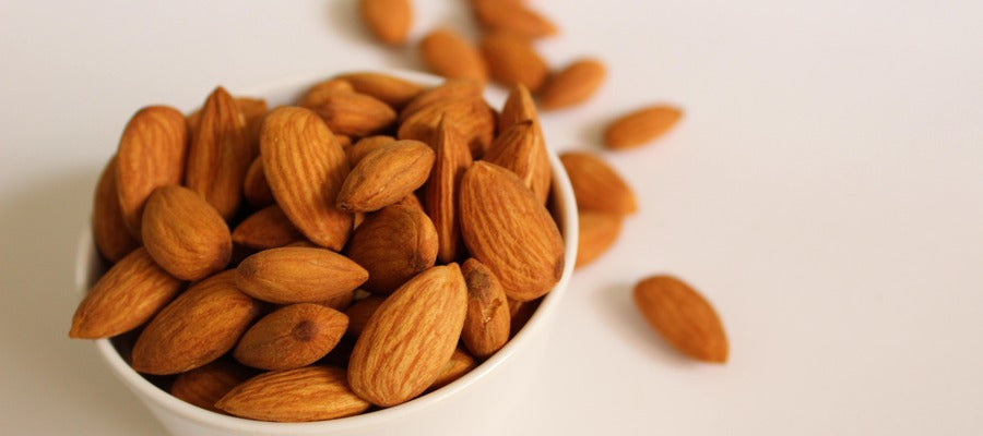 small round bowl of almonds with a few almonds scattered behind on white surface
