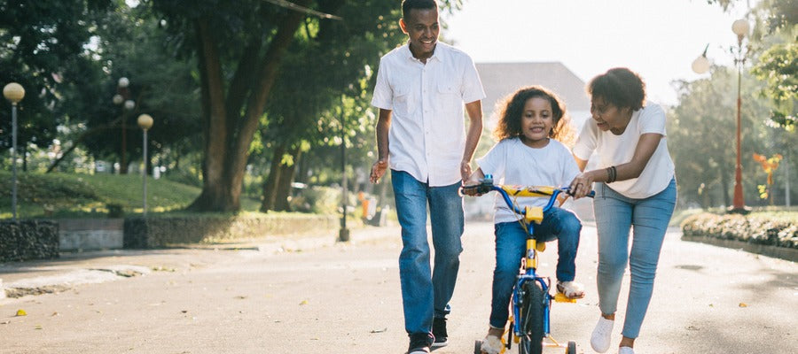 parents helping her young daughter ride a bicycle outdoors on a sunny day with green trees in the background