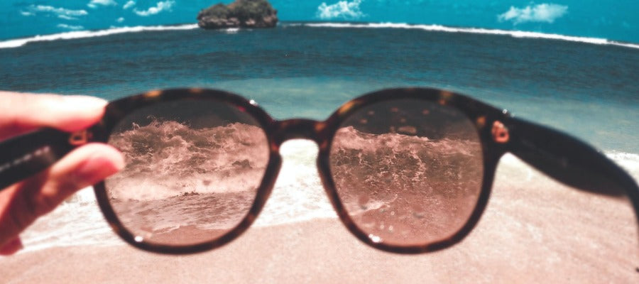 waves and beach seen through a pair of tinted sunglasses held by a hand with azure water and rock in the distance