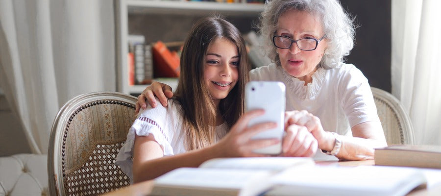 senior woman with white hair and glasses looking into granddaughter's phone at the table with open books before them