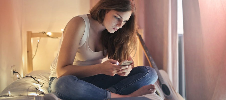 Mujer con fatiga visual sentada con las piernas cruzadas en la cama abotonando su teléfono en su dormitorio