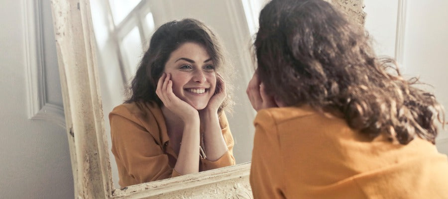 Mujer sonriendo ante su reflejo en un espejo mientras sostiene la barbilla en sus manos.