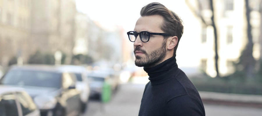 man with dry eyes, glasses, and a black turtleneck looking away against blurry street background