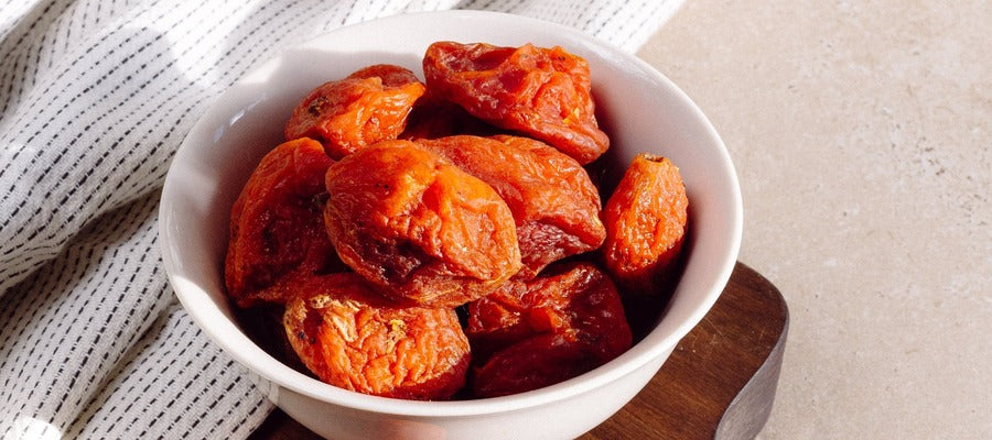 closeup of dried apricots in a white bowl lying on wooden cutter and cloth on table