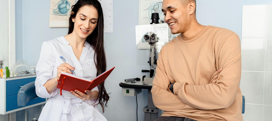 female eye doctor with red notebook smiling and talking with man who sits with arms on chest and with eye exam equipment in the background