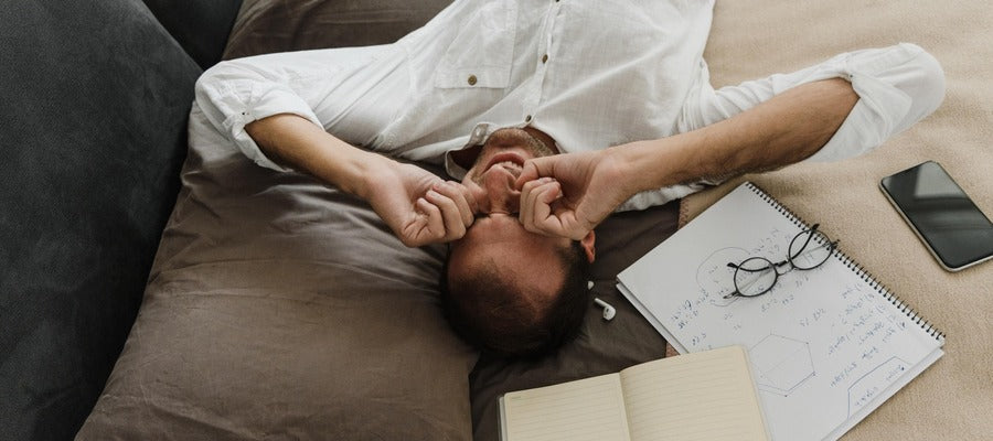 man lying on the bed rubbing his dry eyes with glasses, phone, and notebook to one side seen from above