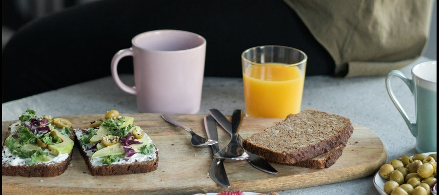 tostada de aguacate sobre un bloque de madera con vajilla, pan integral, vaso de jugo de naranja, café y tazas de té