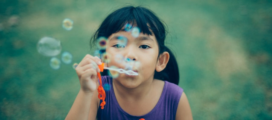 little girl with dark hair and big black eyes blowing soap bubbles against blurry green background