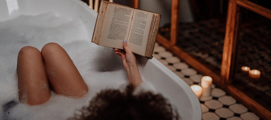 woman reading book in foamy bathtub with candles in the background