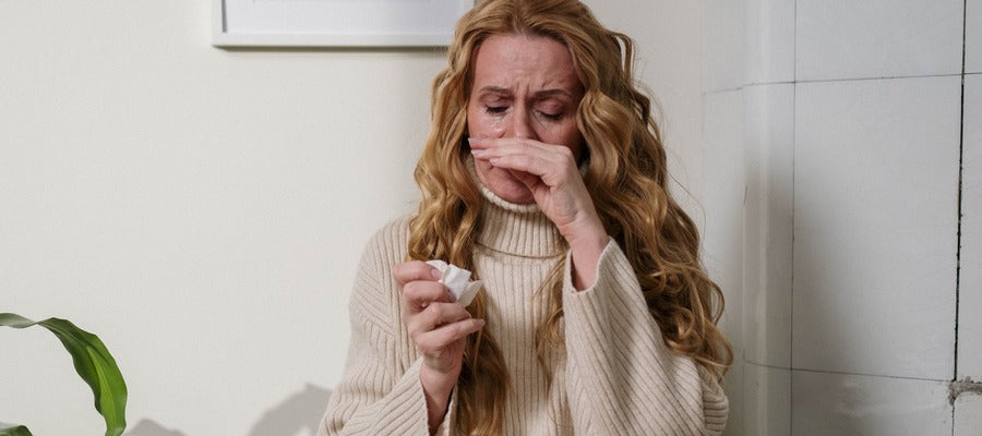 Mujer con cabello rubio largo y ondulado con síntomas de alergia y servilleta en una mano con ojos llorosos y secreción nasal.
