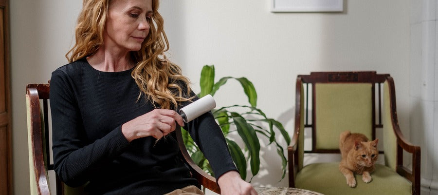 Una mujer con el pelo rubio largo y ondulado cepillando el pelo de su mascota de la manga de su camisa con un rodillo mientras un gato marrón se sienta en la silla al fondo