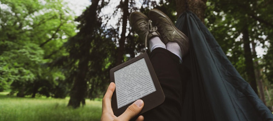 mano sosteniendo un lector de libros electrónicos sobre las piernas estiradas tumbado en una hamaca con árboles y vegetación en el fondo