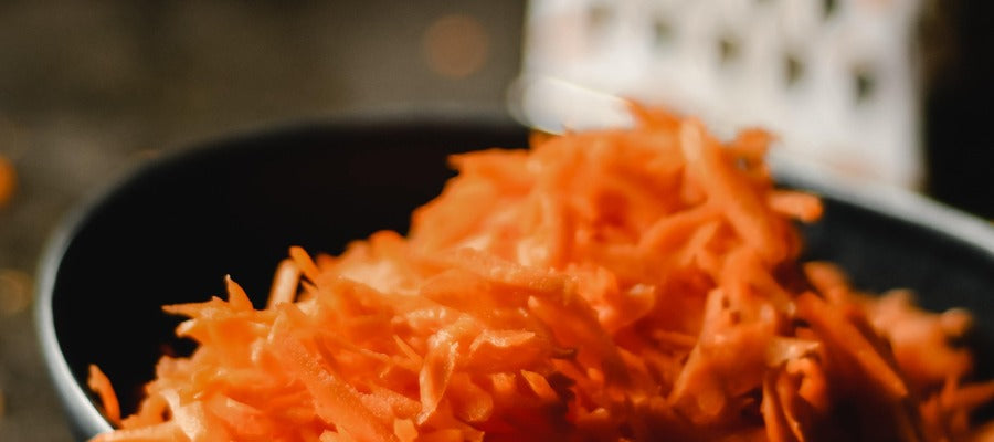 closeup grated carrots in a black bowl with blurry grater in the background