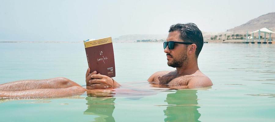 man with sunglasses lying mostly in water at the beach reading a book
